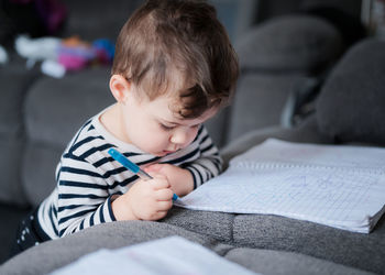 Young baby boy toddler writing in a notebook on the couch at home