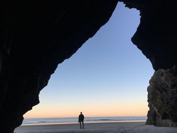 Silhouette man standing by rock formation on beach against sky during sunset