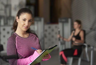 Portrait of fitness coach holding clipboard while woman exercising in background