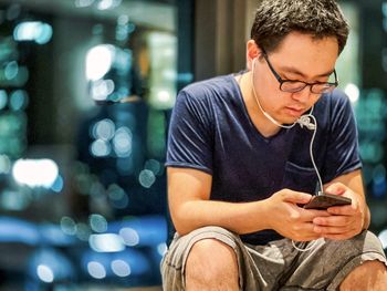 Young asian man with earphones using mobile phone against defocused reflections in window at night.