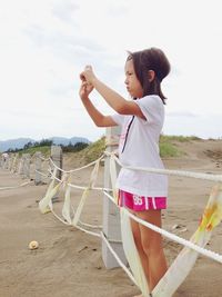 Side view of girl photographing while standing by fence at beach