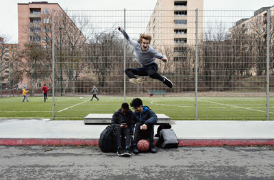 Friends using mobile phone while happy teenage boy jumping on sidewalk in city