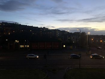 Cars on road by buildings against sky at sunset