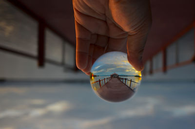 Close-up of person hand holding crystal ball on pier against cloudy sky during sunset