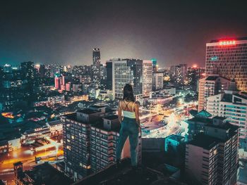 Rear view of woman looking at illuminated cityscape while standing on building terrace against sky