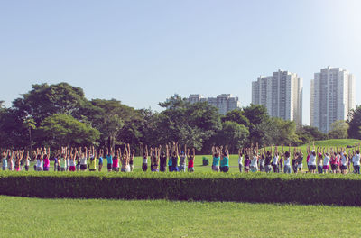 Group of people with arms raised exercising on field against clear sky in city