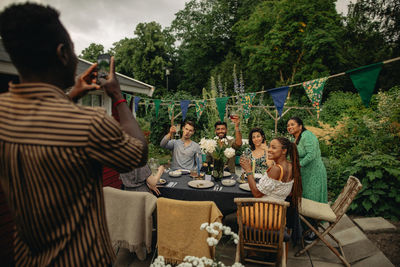 Man photographing male and female friends through smart phone at garden party