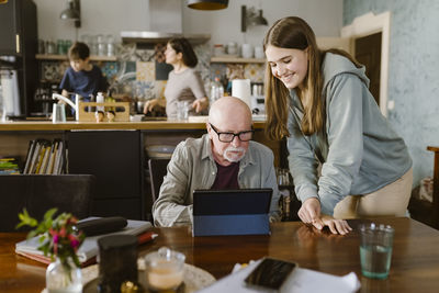 Smiling granddaughter assisting grandfather using tablet pc on table at home