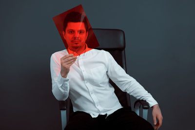 Portrait of young man holding red plastic while sitting on chair against gray background