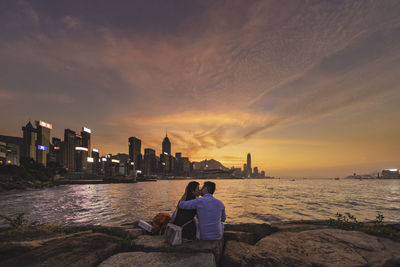 Rear view of people sitting by sea against sky during sunset