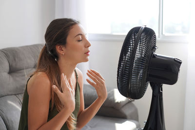 Young hispanic woman cooling down by ventilator at home, feeling unwell with high temperature 