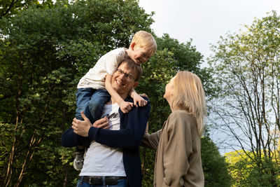 Father carried little child, son on shoulder. mother walks beside in park. summer time. happy