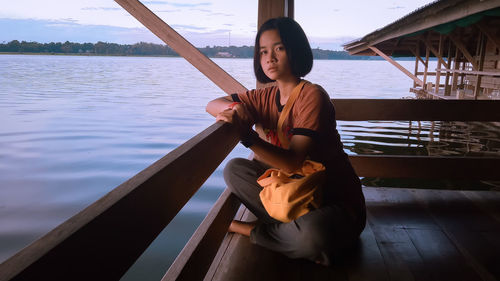 Portrait of young woman sitting on stilt house over lake during sunset