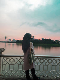 Rear view of woman standing by railing against river during sunset