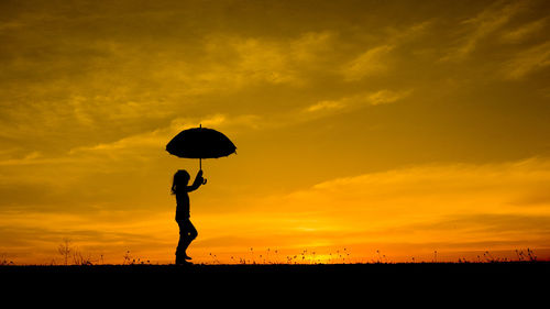 Silhouette girl with umbrella walking on field against sky during sunset