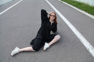 Fashion portrait of ginger woman in loose black dress and sneakers sitting posing on road outside 