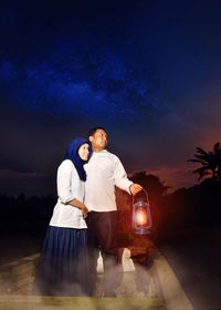 Couple with oil lamp standing on boat against sky at night