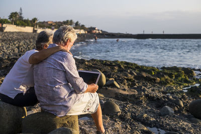 Senior man with woman using laptop while sitting on rock at beach