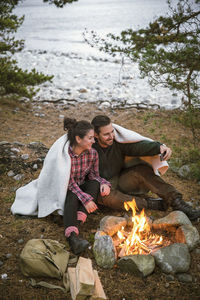 Couple wrapped in blanket taking selfie while sitting by fire pit at campsite