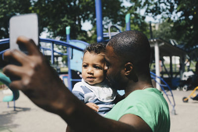 Father kissing toddler while taking selfie through mobile phone at park