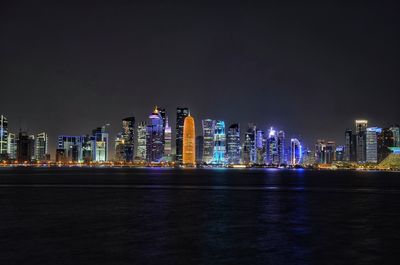 Illuminated buildings by sea against sky at night