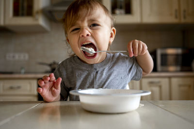 Baby boy eating porridge with spoon in kitchen