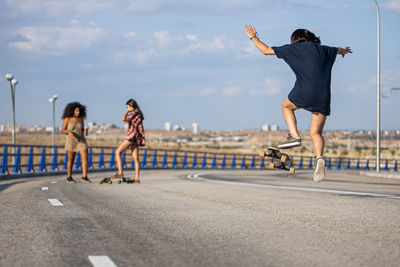Unrecognizable young woman doing a trick with her long board by a bridge with her companions in the background