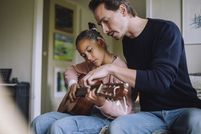 Father assisting daughter playing guitar while sitting on bed at home