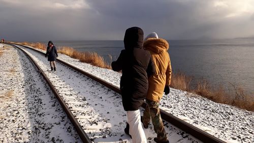 Rear view of people walking on snow covered railroad track by sea