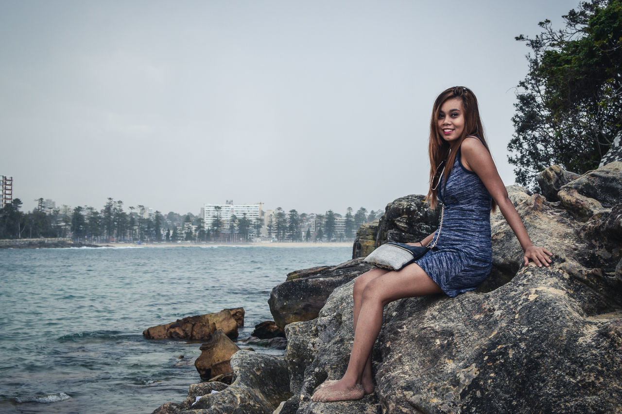 sitting, adults only, young adult, portrait, city, water, looking at camera, outdoors, day, adult, people, only women, happiness, one person, full length, sky, young women, beautiful woman, smiling, one woman only, urban skyline, architecture, one young woman only, millionnaire