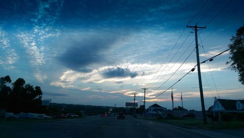 Electricity pylons on road against cloudy sky