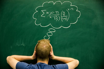 Rear view of man with head in hands standing by formula on blackboard