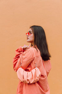 Stylish female in trendy outfit and matching handbag and sunglasses standing near orange wall