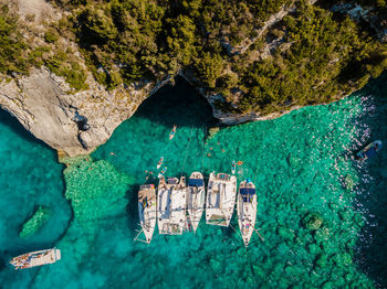 Aerial view of sea cave and rock formation with sailboats anchored and group swimming in blue water.
