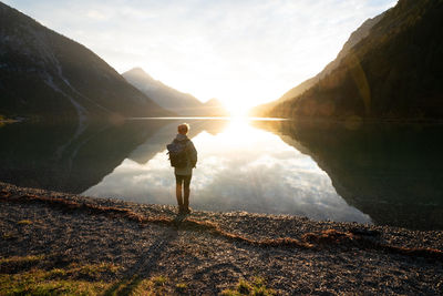 Rear view of man standing by lake amidst mountains during sunny day