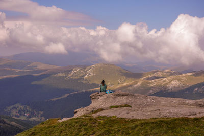 Woman sitting on cliff against cloudy sky during sunny day