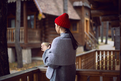 Woman in a red hat and scarf and a mug stands at a wooden house, in the woods in the autumn morning 