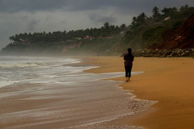 Rear view of woman walking at beach against cloudy sky