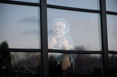 Sad mature woman with scraft on head standing behind dirty window glass