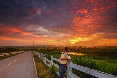 Man leaning on railing by road against sky during sunset