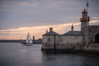 Sailboats on sea by buildings against sky during sunset