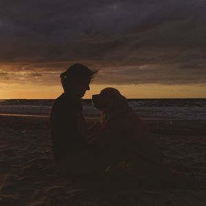 Woman sitting with dog at beach against sky during sunset