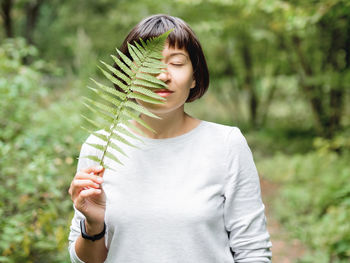 Young woman is hiding her eye with fern leaf. symbol of lifeand unity with nature. summer in forest.