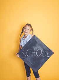 Portrait of smiling girl holding blackboard with school text while standing against yellow background