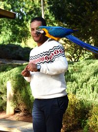 Man with macaw on shoulder standing against plants in park