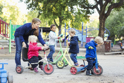 Female teacher assisting girl to ride tricycle on playground