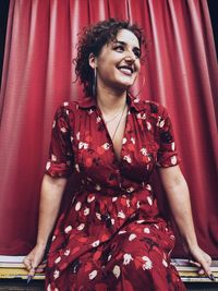 Smiling woman looking away sitting against curtain