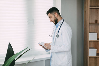 Modern young male doctor stands in his office and writes in his notebook