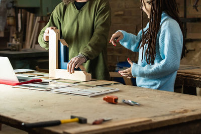 Crop carpenter standing at workbench with wooden detail and tools with laptop while discussing order with customer