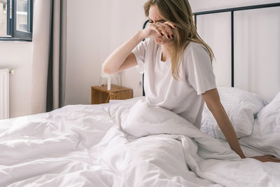 Sleepy young woman rubs her eyes while sitting in bed in the morning feeling tired after waking up. 